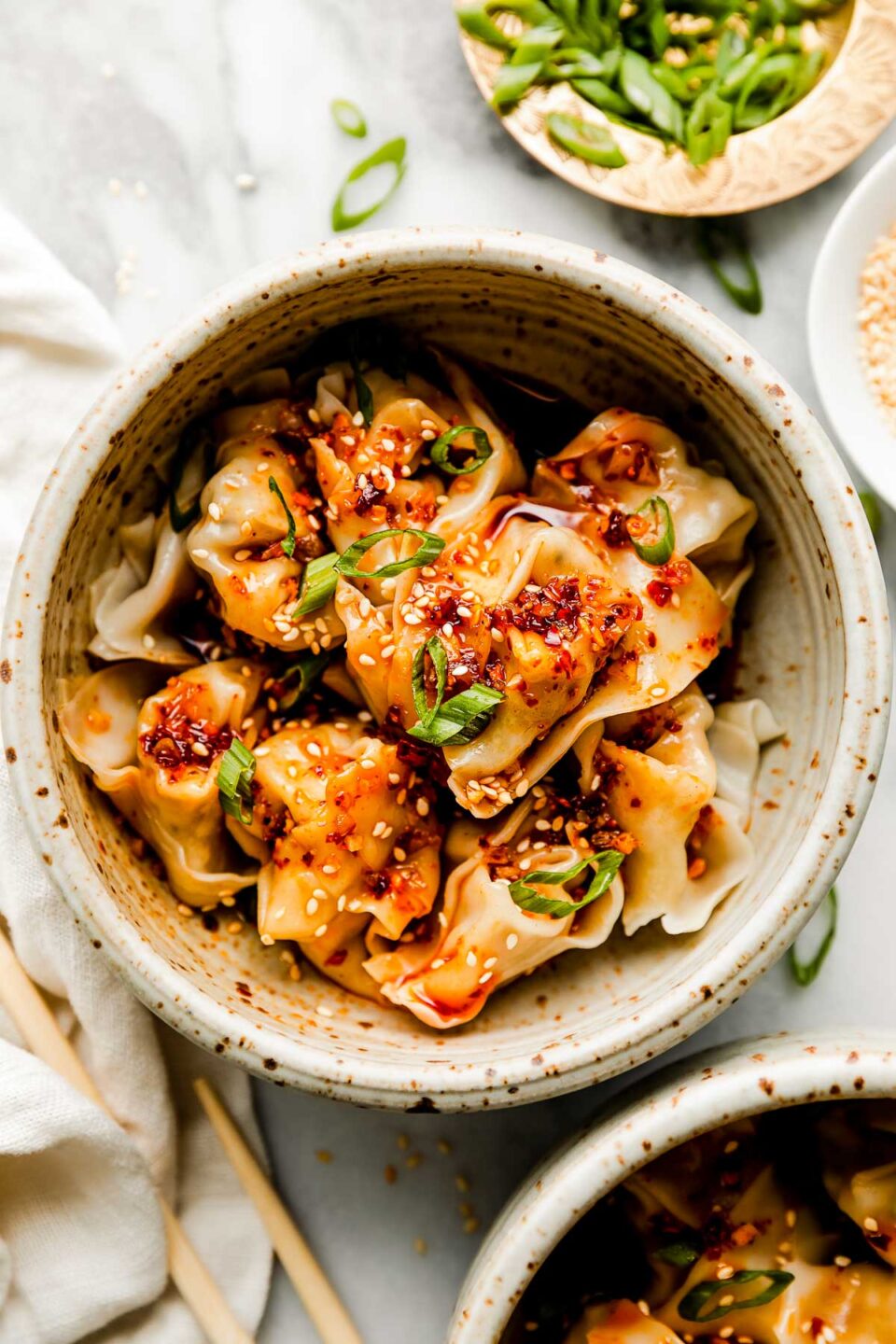An overhead shot of a stoneware bowl of cooked wontons, topped with spicy chili oil and sliced green onions atop a white marbled surface alongside chopsticks and a white dish towel. Smaller dishes of sliced green onions, spicy chili oil, and sesame seeds sit beside it.
