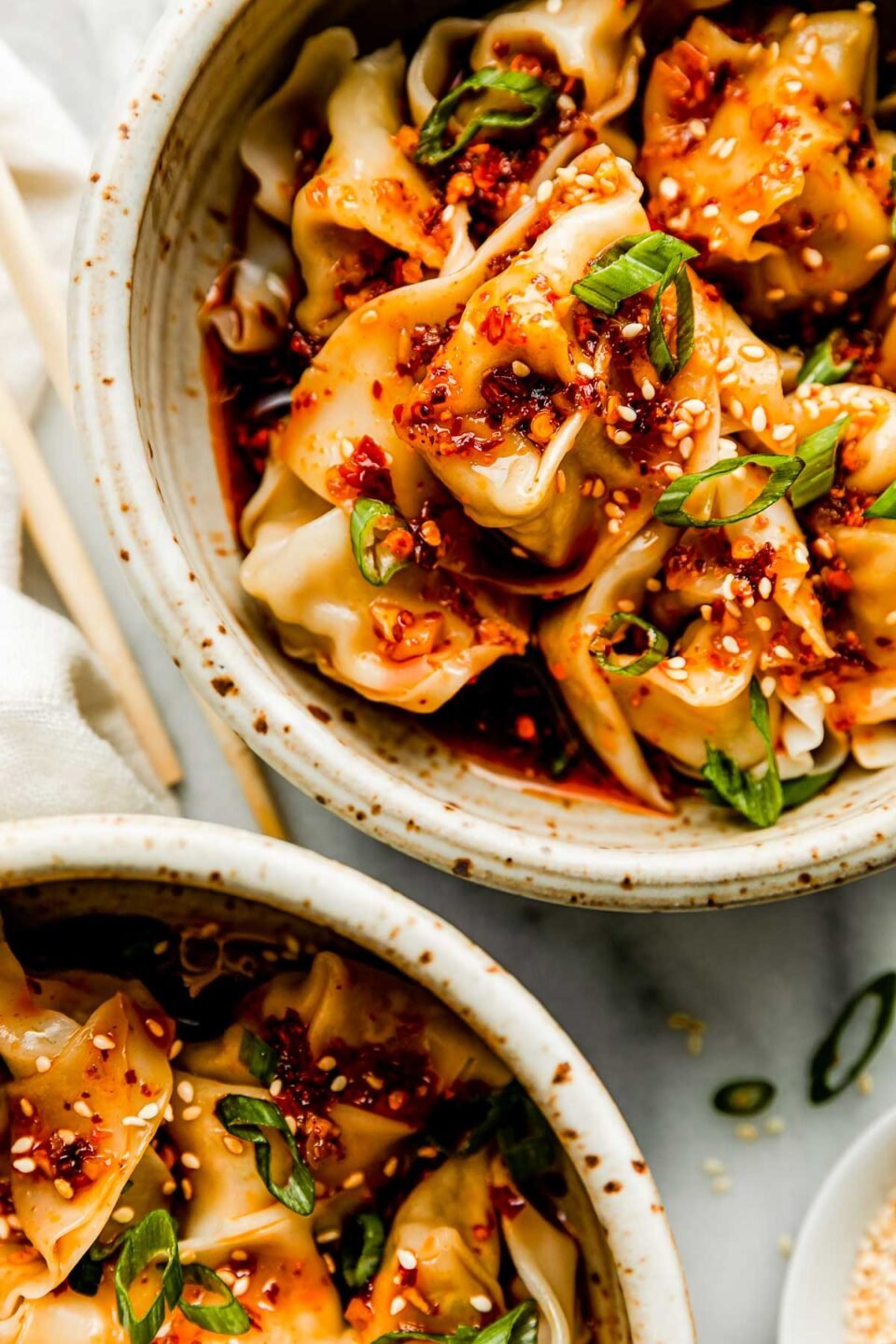 A close-up overhead shot of 2 bowls of cooked wontons topped with spicy chili oil, sesame seeds & green onions atop a white marbled surface. chopsticks, a white dish towel and small dish of sesame seeds surround the bowls.