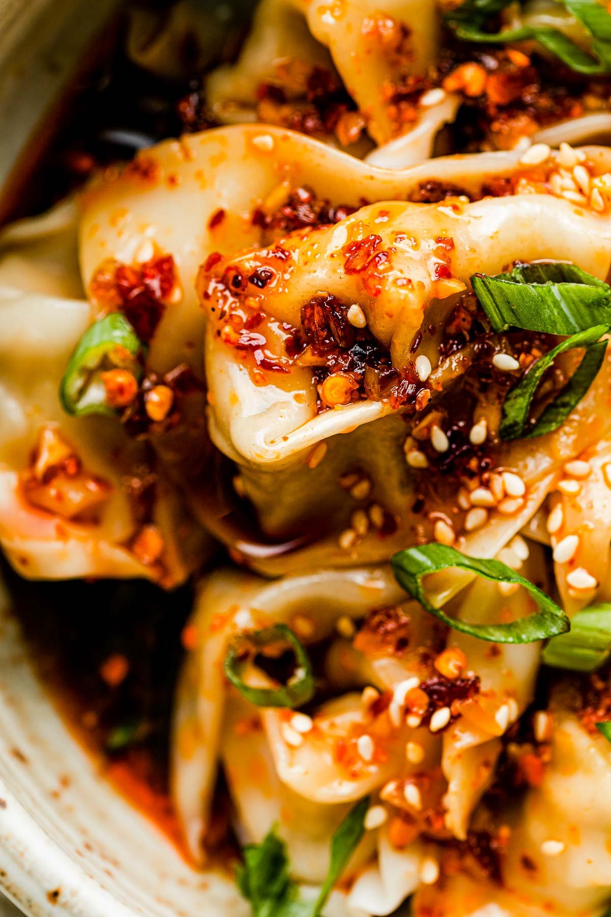 A close-up overhead shot of a bowl of cooked wontons topped with spicy chili oil, sesame seeds & green onions atop a white marbled surface. A wonton is being picked up by a pair of bamboo chopsticks.