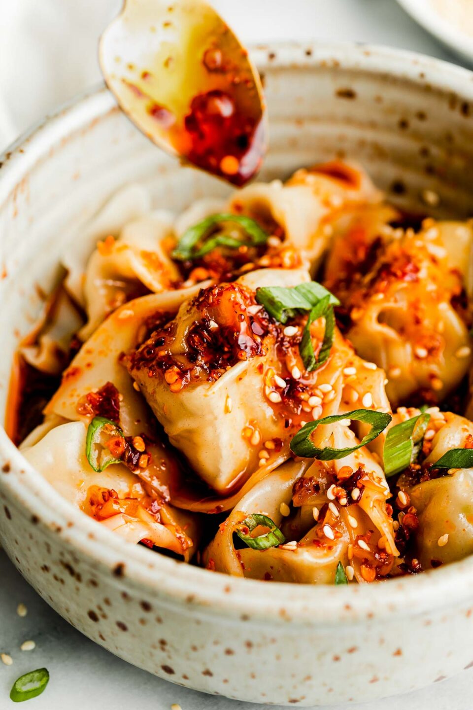 A close-up side shot of cooked wontons coated with spicy chili oil, sesame seeds and sliced green onions in a stoneware bowl. A gold spoon is drizzling spicy chili oil over the top.