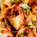A close-up macro shot of cooked wontons coated with spicy chili oil, sesame seeds and sliced green onions.