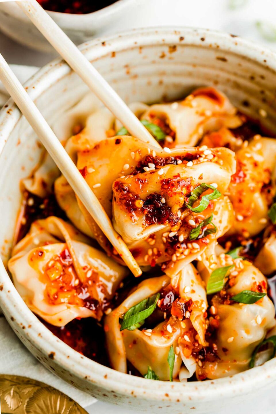 A close-up overhead shot of a bowl of cooked wontons topped with spicy chili oil, sesame seeds & green onions atop a white marbled surface. A wonton is being picked up by a pair of bamboo chopsticks.