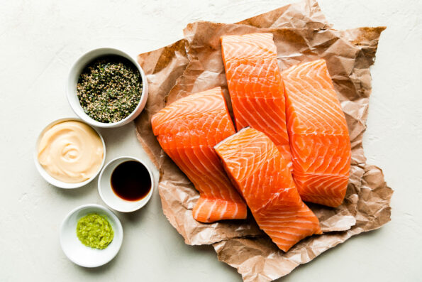 Furikake Salmon ingredients sit on a textured white surface: raw salmon fillets on brown butcher paper, and small bowls of furikake, shoyu, wasabi & Kewpie mayonnaise.