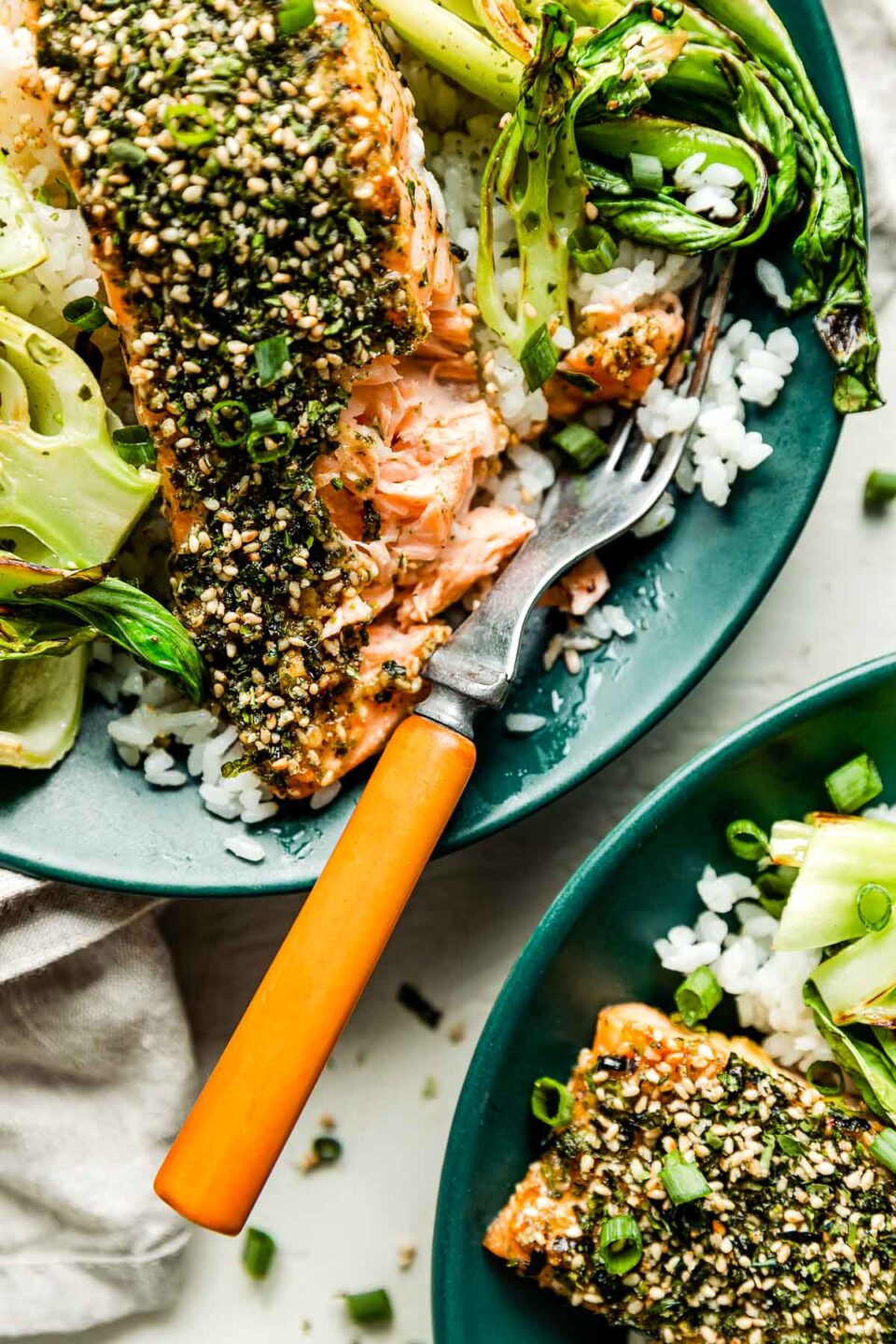 A close-up overhead shot of the cooked furikake salmon on a bed of rice with bok choy in a dark green bowl with a wood-handled fork. A second bowl of prepared food sits beside it.
