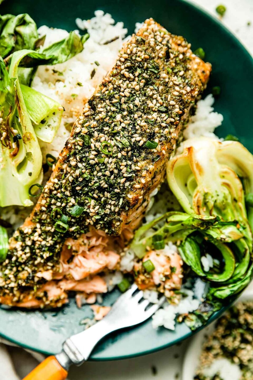 A close-up overhead macro shot of a partially eaten furikake salmon fillet on a green plate with bok choy, white rice & a wood-handled fork.