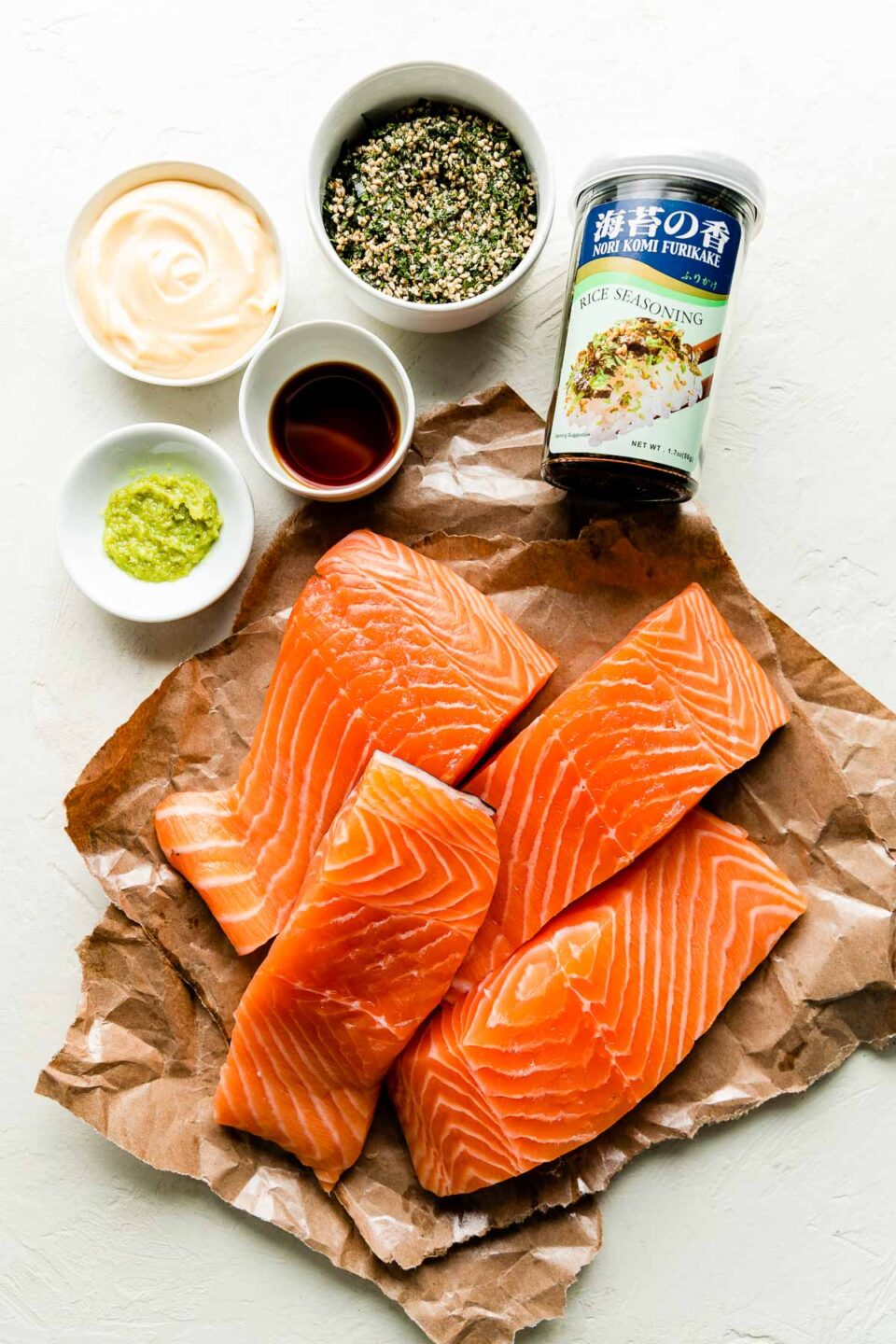 An overhead shot of ingredients sitting on a textured white surface: raw salmon fillets on brown butcher paper, a small tin of furikake, and small bowls of furikake, shoyu, wasabi & Kewpie mayonnaise.