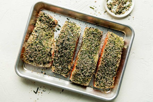 An overhead shot of the seasoned furikake salmon ready to be baked on a sheer pan atop a white surface. A small dish of furikake sits beside it.
