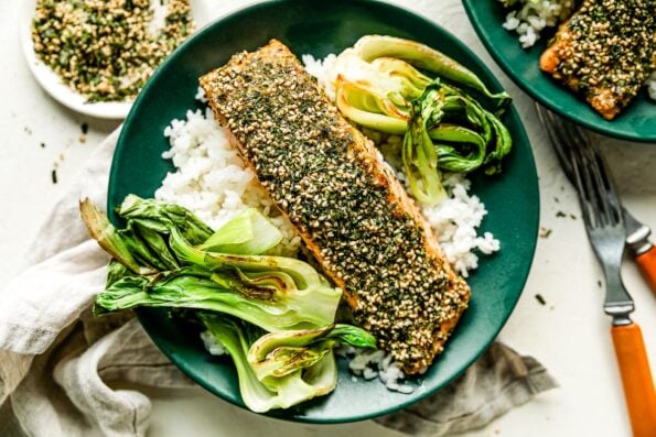 A green shallow bowl with cooked white rice, furikake salmon and bok choy sits on a dishtowel on a white surface alongside a wood-handled fork and knife, dish of furikake seasoning and another prepared bowl.