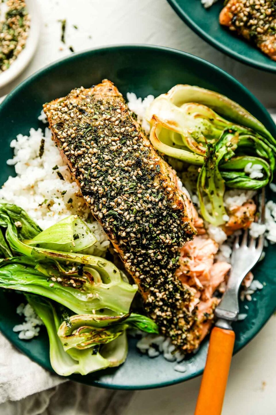 A green shallow bowl with cooked white rice, furikake salmon and bok choy sits on a dishtowel on a white surface alongside a wood-handled fork, dish of furikake seasoning and another prepared bowl.