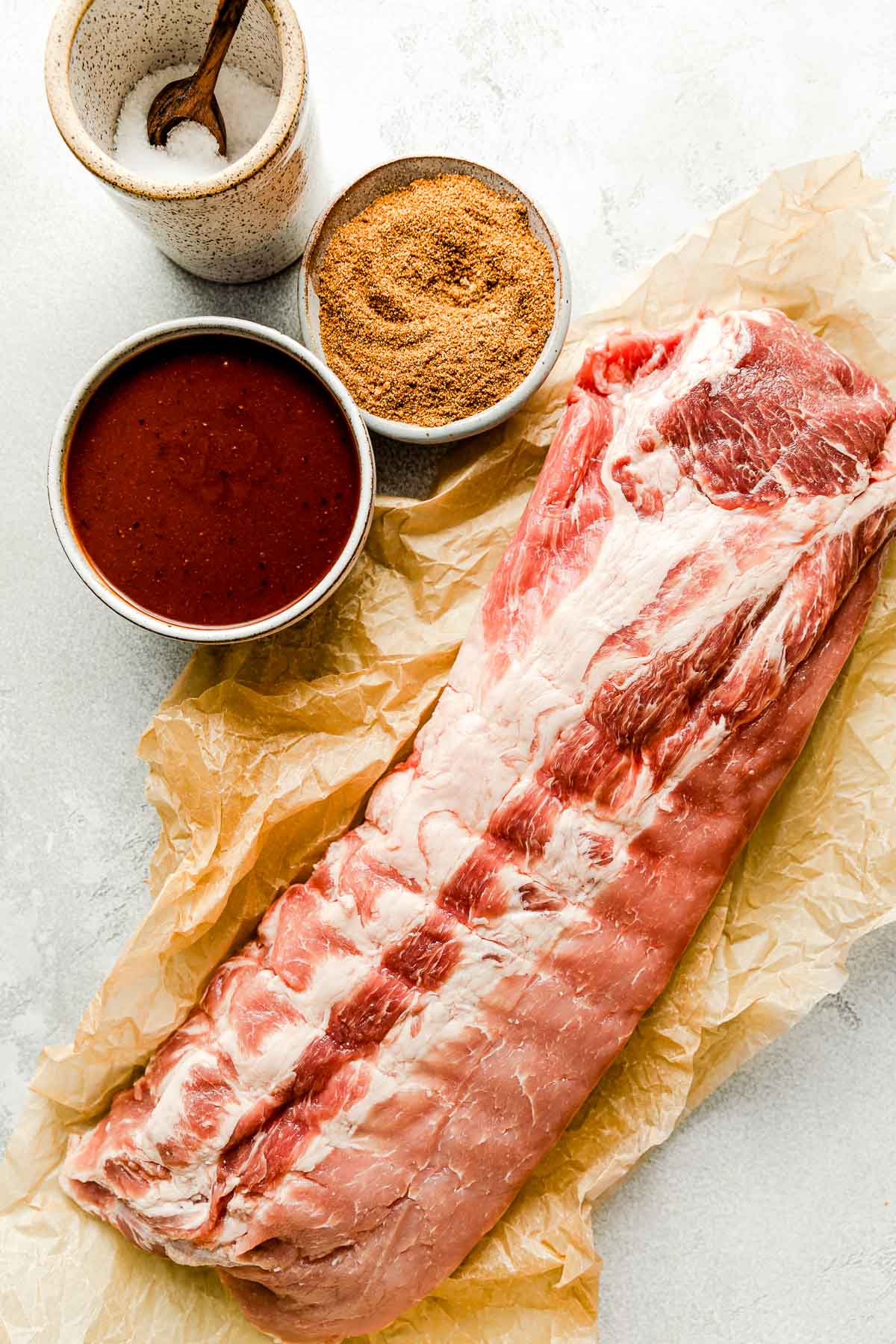 Grilled baby back ribs ingredients sit atop a white marbled surface: dry rub, A macro close-up shot from the side of charred baby back ribs on the grill, being brushed with BBQ sauce. sauce, kosher salt & baby back ribs,