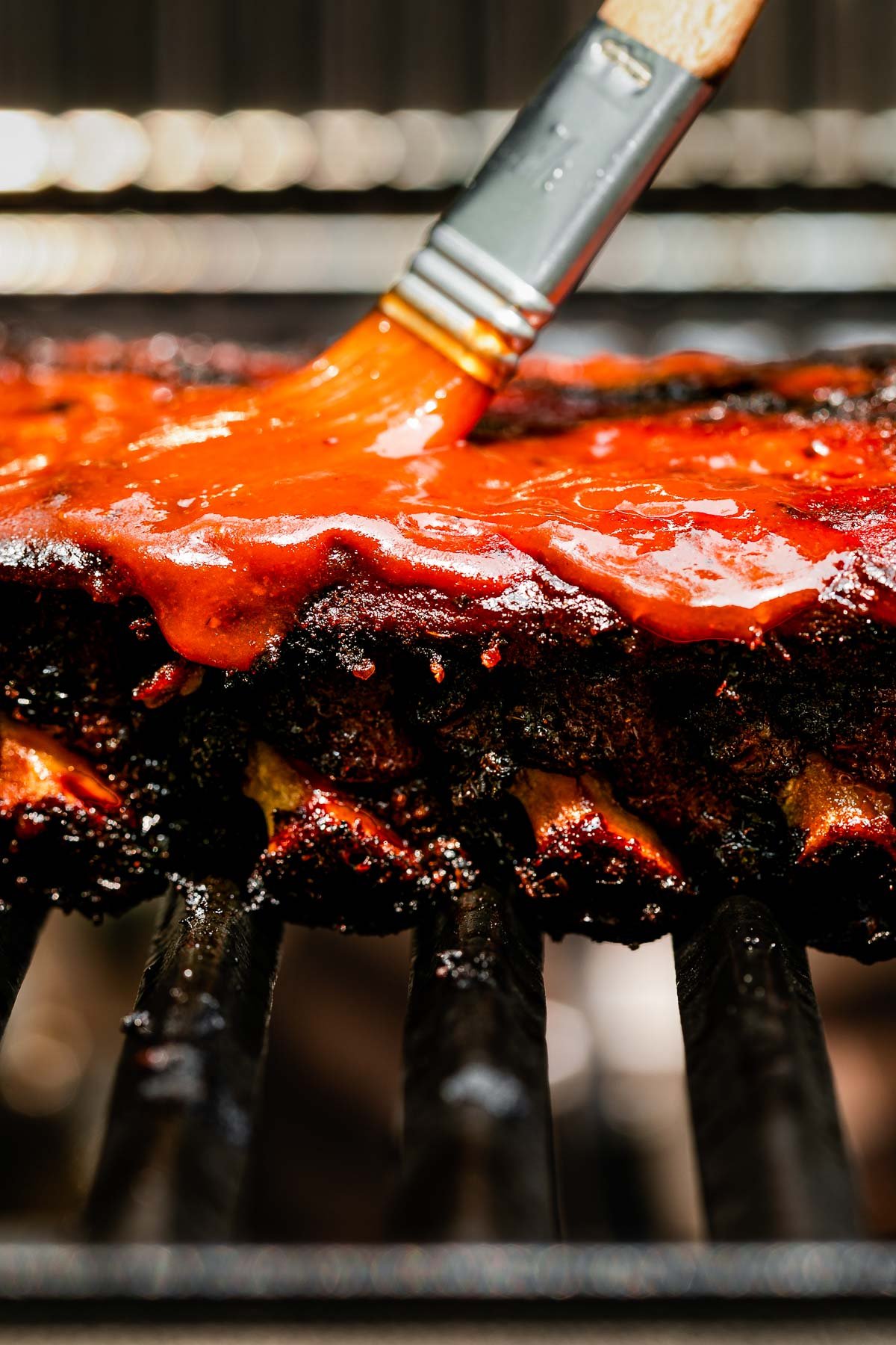 A macro close-up shot from the side of charred baby back ribs on the grill, being brushed with BBQ sauce.