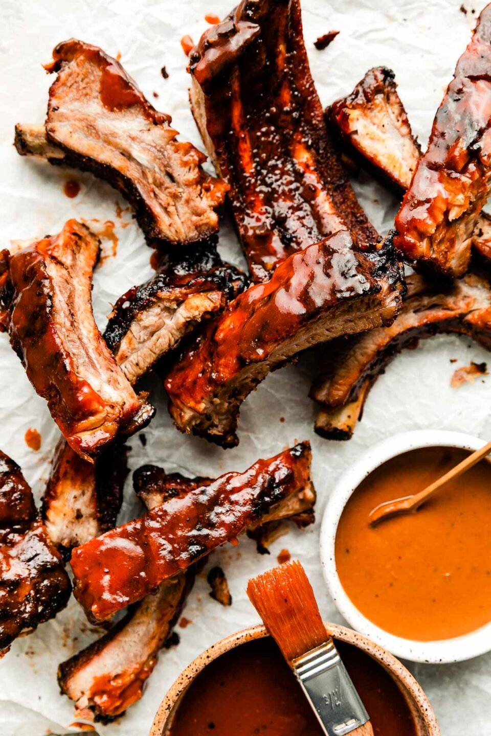 An overhead shot of individual grilled ribs on a white surface. Two bowls of BBQ sauce and a pastry brush sit alongside them.