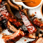 A close-up overhead shot of individual grilled ribs on a white surface. Two bowls of BBQ sauce sit alongside them.