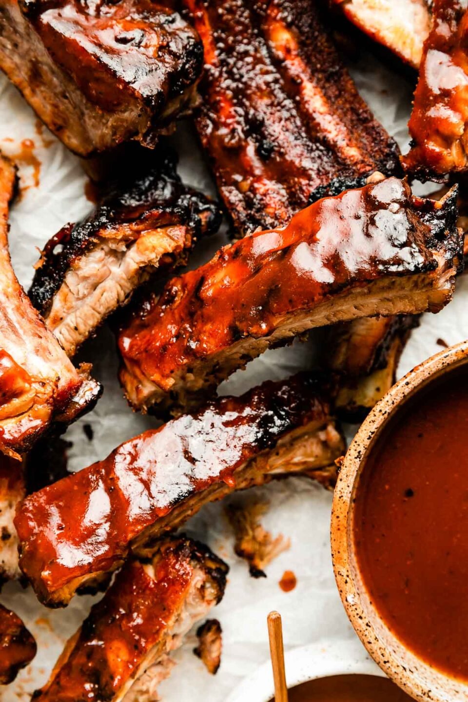 An overhead shot of individual grilled ribs atop a white surface. A bowl of BBQ sauce sits alongside the ribs.