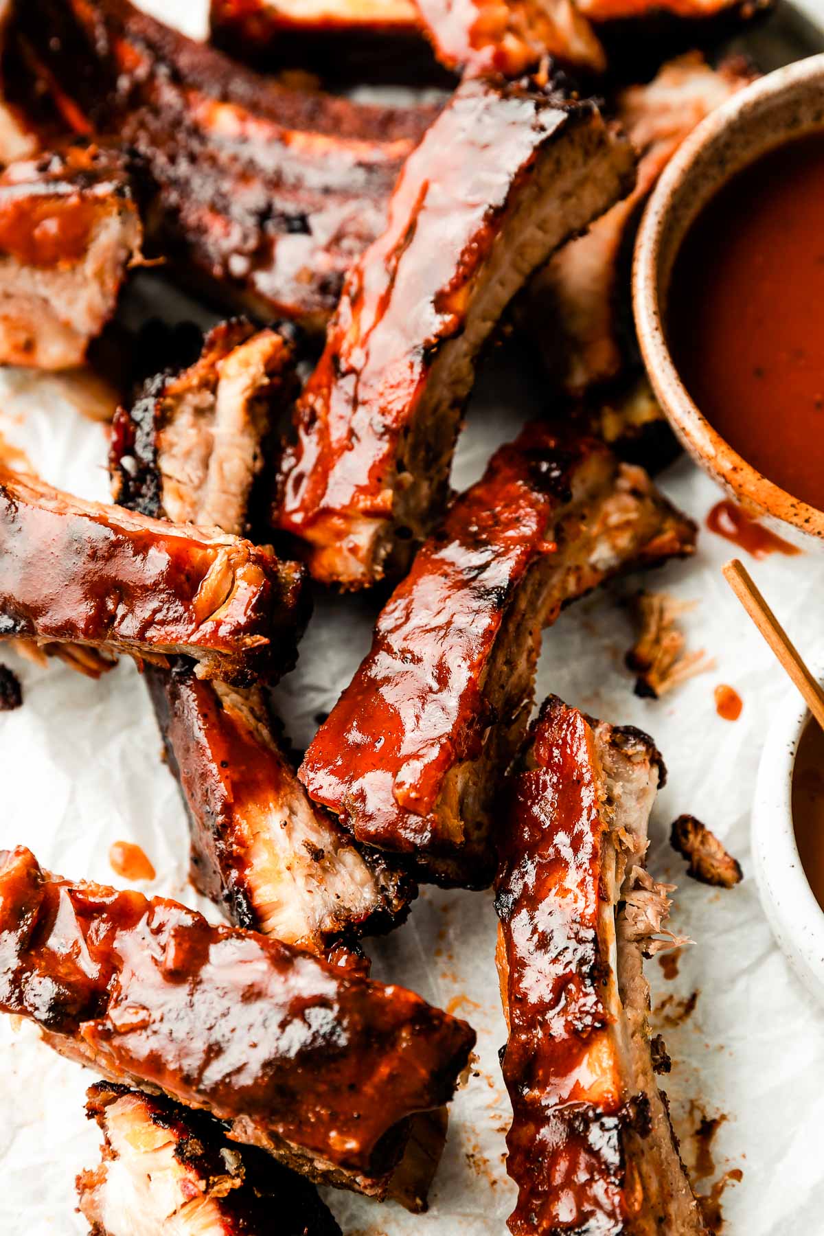 An overhead shot of individual grilled ribs on a paper-lined sheet pan. A bowl of BBQ sauce sits alongside them.