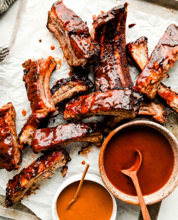 An overhead shot of individual grilled ribs on a paper-lined sheet pan atop a white surface. Two bowls of BBQ sauce sit alongside them.