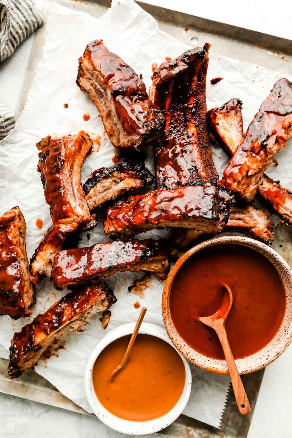 An overhead shot of individual grilled ribs on a paper-lined sheet pan atop a white surface. Two bowls of BBQ sauce sit alongside them.