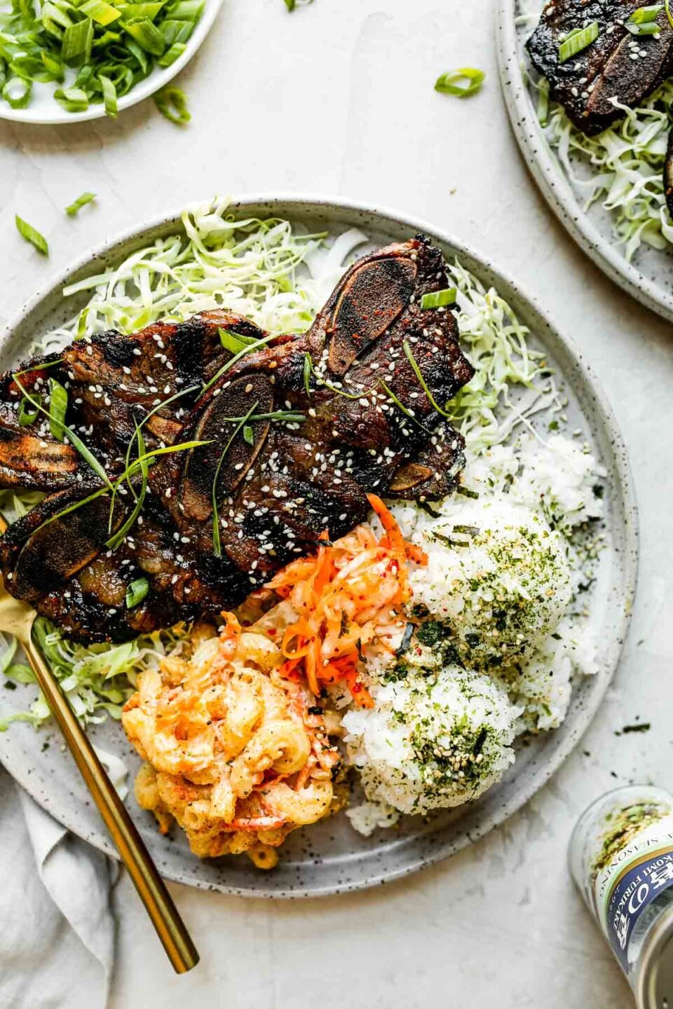 Kalbi served local Hawaiian-style, plated with shredded cabbage & scoops of rice, mac salad, & kimchi. The plate sits atop a cement surface alongside a second plate of kalbi, nori furikake seasoning, a gray linen napkin, & a plate of thin sliced green onions.