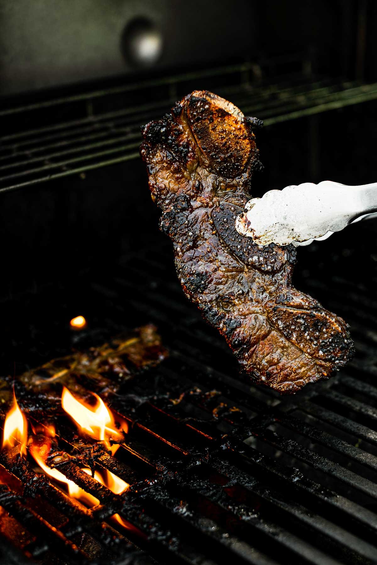 Grill tongs holding grilled Korean short ribs over grill grates. The kalbi is charred & caramelized.