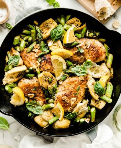 An overhead shot of the baked lemon chicken skillet atop a white marbled surface, with browned chicken breast, chopped asparagus, artichoke hearts and garlic. The skillet is topped with fresh basil leaves and lemon wedges, and surrounded by glasses of white wine and a loaf of crusty bread on a wooden cutting board.