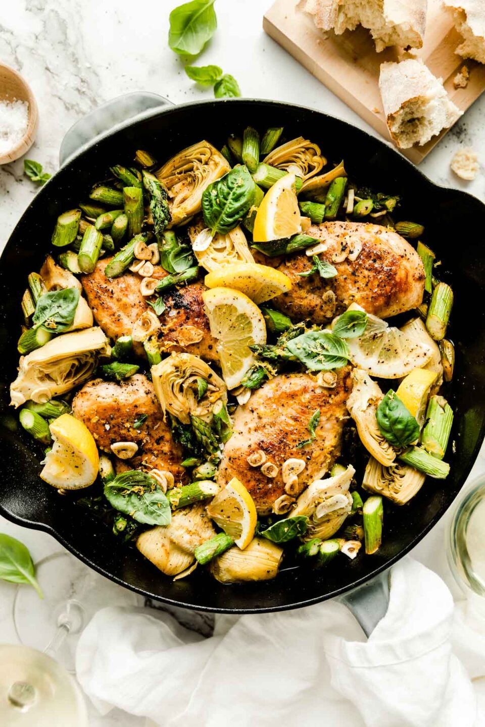 An overhead shot of the baked lemon chicken skillet atop a white marbled surface, with browned chicken breast, chopped asparagus, artichoke hearts and garlic. The skillet is topped with fresh basil leaves and lemon wedges, and surrounded by glasses of white wine and a loaf of crusty bread on a wooden cutting board.