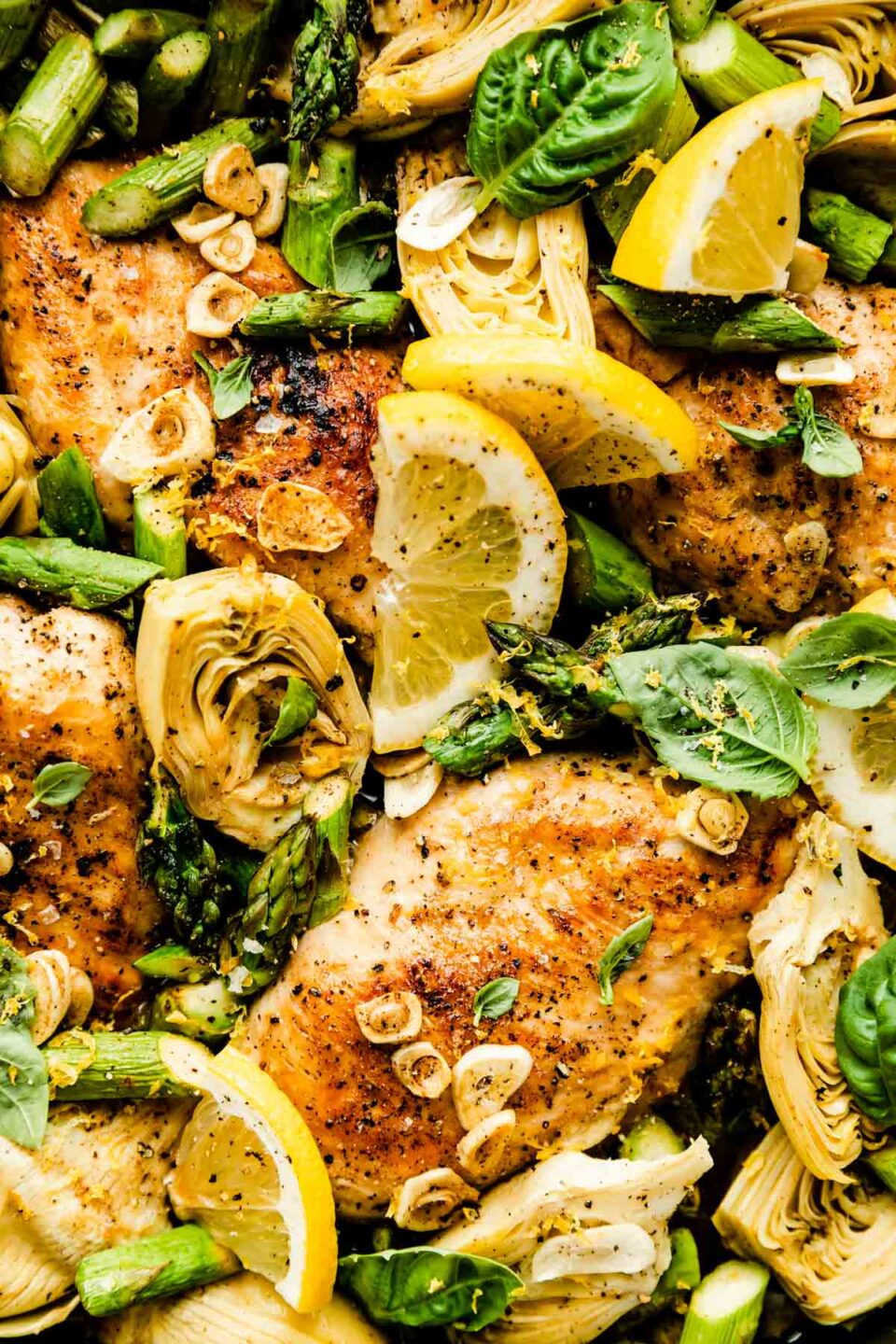 A close-up overhead shot of the baked lemon chicken skillet, with browned chicken breast, chopped asparagus, artichoke hearts and garlic. The skillet is topped with fresh basil leaves and lemon wedges.