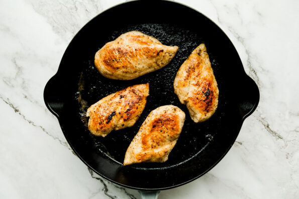 An overhead shot of browned chicken breasts in a black skillet sitting atop a white marbled surface.