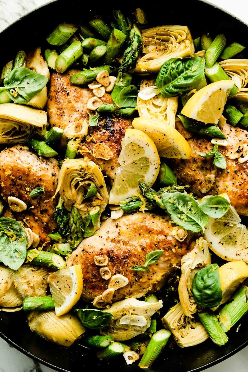 An overhead shot of the baked lemon chicken skillet, with browned chicken breast, chopped asparagus, artichoke hearts and garlic. The skillet is topped with fresh basil leaves and lemon wedges.