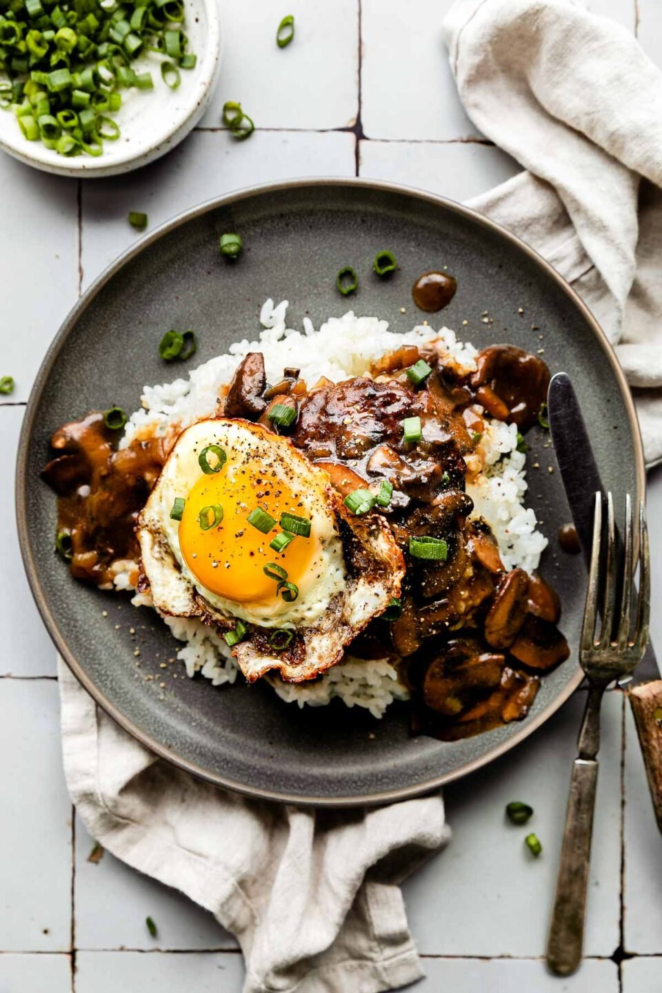 An overhead shot of loco moco served on a bed of rice on a sage green plate atop a grey tiled surface, with a white dishtowel, wood-handled fork & knife, and dish of green onions surrounding it.