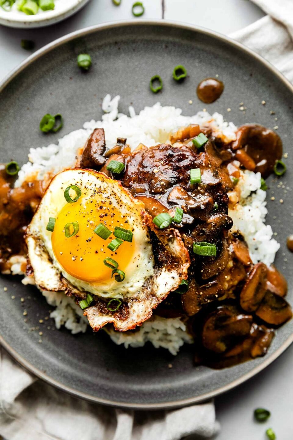 A close-up overhead shot of loco moco served on a bed of rice on a sage green plate atop a grey tiled surface, with a white dishtowel and dish of green onions surrounding it.