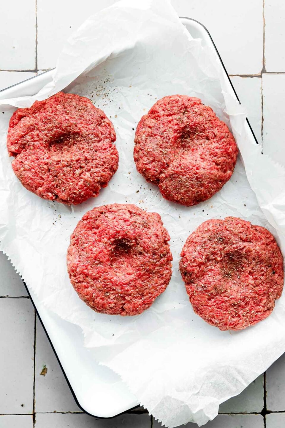 4 raw beef patties, each with an indent in the middle, rest on a parchment paper-lined sheet pan atop a grey tiled surface.