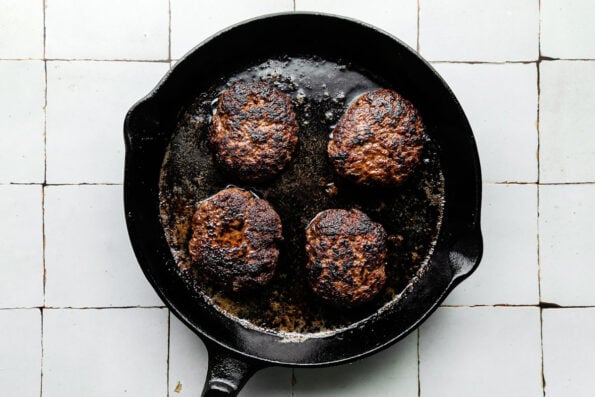 Four cooked beef patties sit in a black cast-iron skillet atop a white-grey tiled surface.