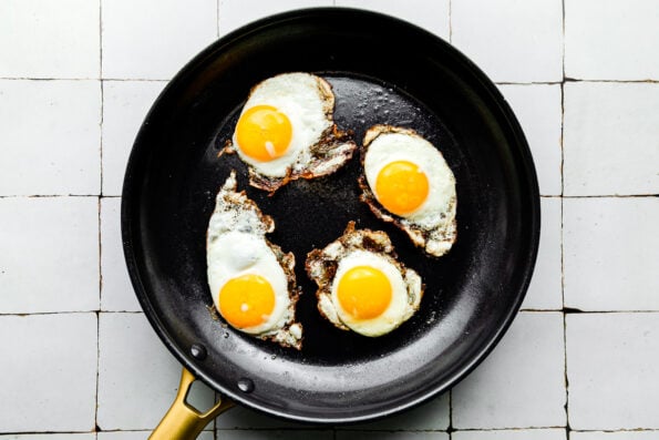 Four sunny-side-up fried eggs sit in a black skillet with a gold handle atop a white-grey tiled surface.
