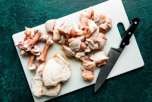 Chicken thighs, some cut and some whole, sit on a white cutting board with a butcher's knife, atop a teal textured surface.