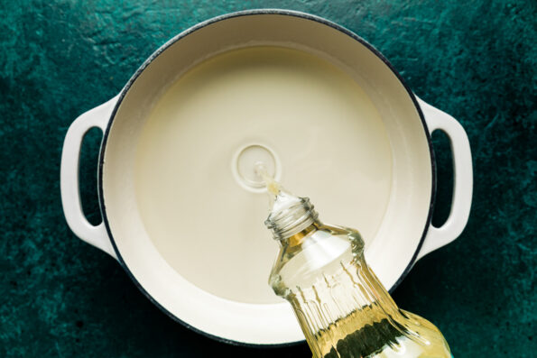 Overhead shot of oil being poured into a white Dutch oven atop a dark teal textured surface.