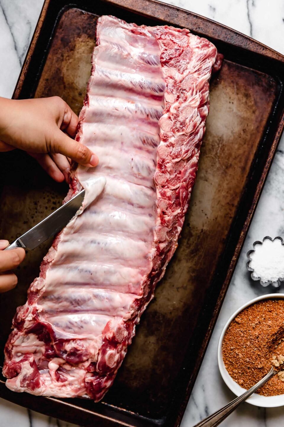 An overhead shot of raw baby back ribs on a sheet pan atop a white marbled surface, with a woman's hand holding a knife & removing the silverback membrane from the ribs. A small dish of dry rub and a small dish of salt sit alongside the pan.