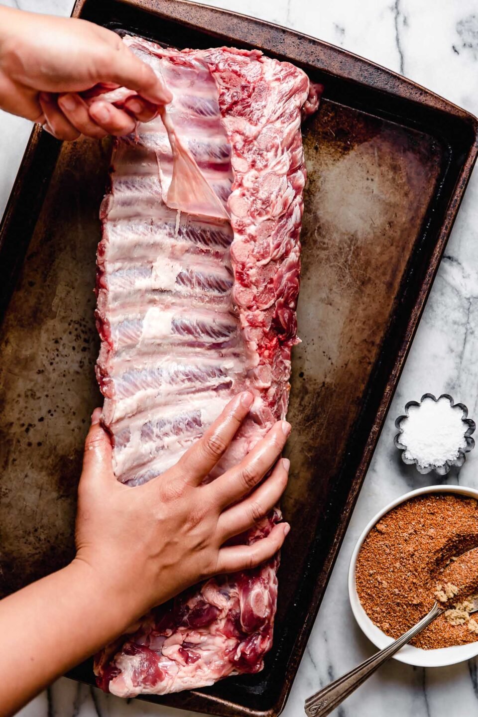 An overhead shot of raw baby back ribs on a sheet pan atop a white marbled surface, with a woman's hand removing the silverback membrane from the ribs. A small dish of dry rub and a small dish of salt sit alongside the pan.