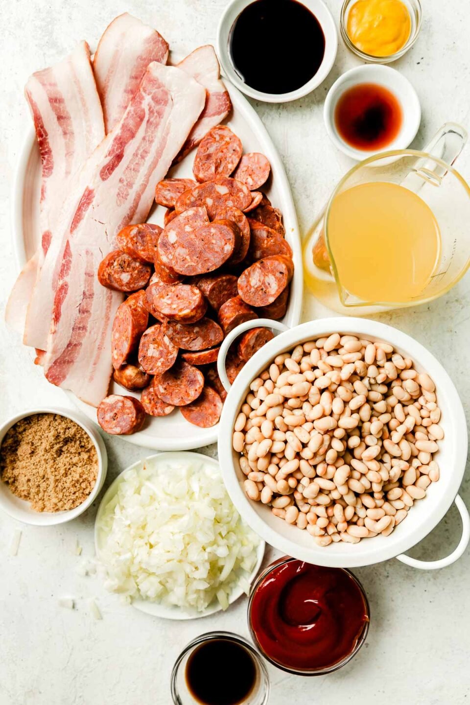 Brown sugar baked beans ingredients sit atop a white textured surface: beans, sliced sausage, raw bacon, chopped onions and sauce ingredients.