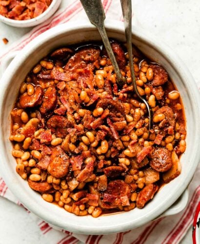 A white crock full of baked beans sits atop a red and white striped cloth on a white textured surface. A small dish of crumbled bacon sits next to it.