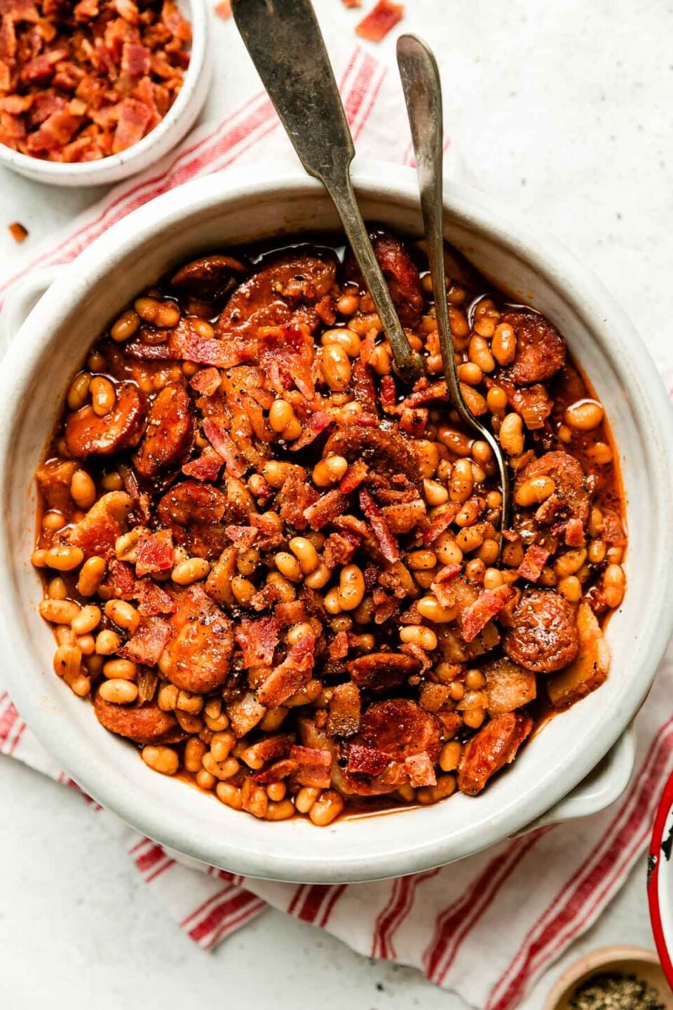 A white crock full of baked beans sits atop a red and white striped cloth on a white textured surface. A small dish of crumbled bacon sits next to it.