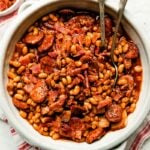 A white crock full of baked beans sits atop a red and white striped cloth on a white textured surface. Small dishes of crumbled bacon and ground black pepper sit next to it.