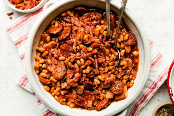 A white crock full of brown sugar baked beans sits atop a red and white striped cloth on a white textured surface. Small dishes of crumbled bacon and ground black pepper sit next to it.