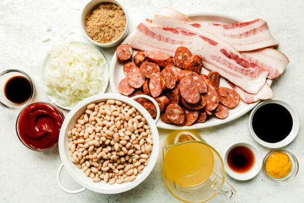 Brown sugar baked beans ingredients sit atop a white textured surface: beans, sliced sausage, raw bacon, chopped onions and sauce ingredients.