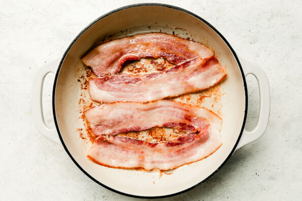 A white skillet with rendered bacon slices sits atop a white textured surface.