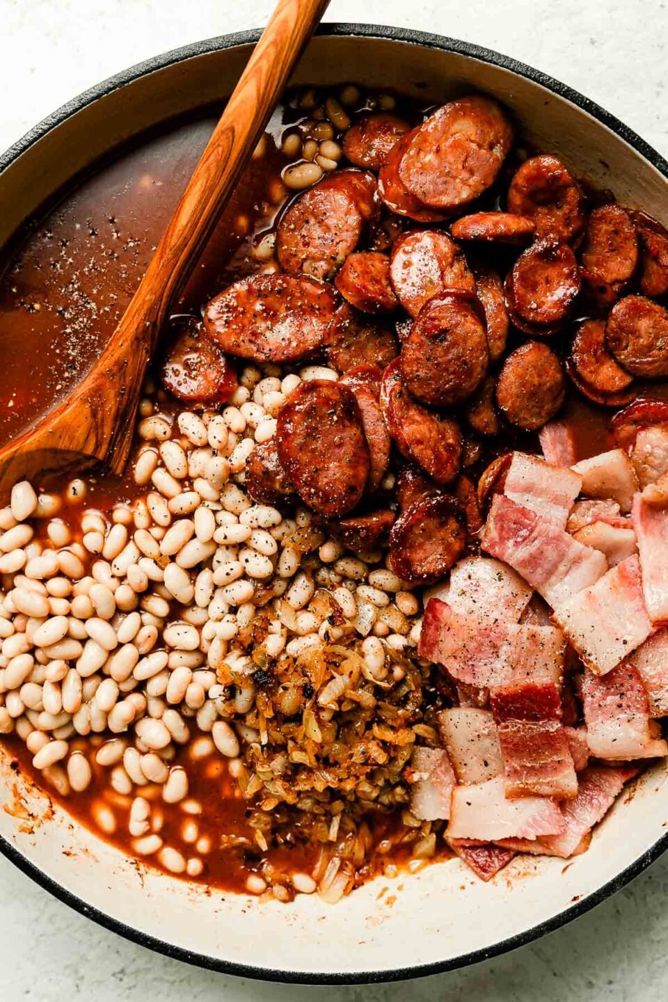 Prepared brown sugar baked beans ingredients sit in a white skillet atop a white surface: rendered bacon, cooked sausage, sauce, beans & onions.