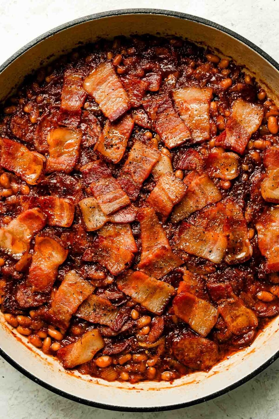 A white skillet of baked brown sugar baked beans with bacon and sausage sits atop a white textured surface.