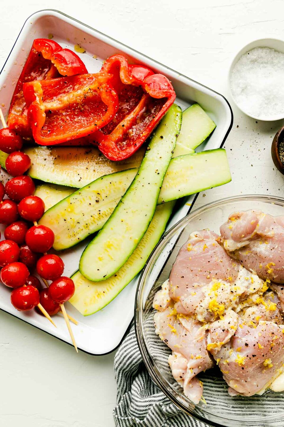 A baking dish with ready-to-grill zucchini, red peppers and skewers of tomatoes, and a bowl of marinated chicken thighs sit atop a white surface. Small bowls of salt and pepper sit alongside them.