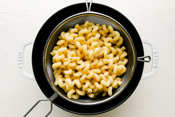 An overhead shot of cooked noodles in a silver strainer resting in a cast iron pot atop a white surface.
