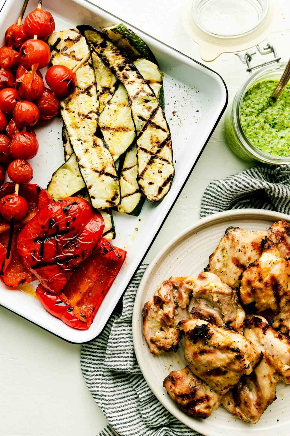 An overhead shot of a baking dish with grilled tomato skewers, red peppers, and zucchini, and a plate of grilled chicken atop a striped towel on a white surface. A jar of pesto sits beside them.