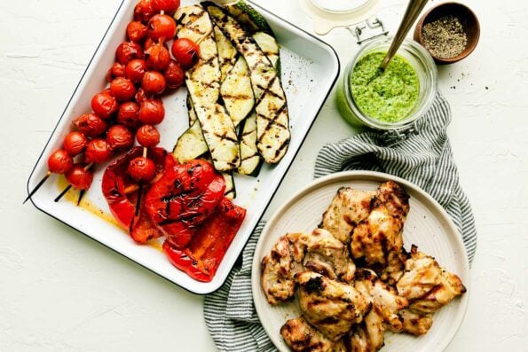 An overhead shot of a baking dish with grilled tomato skewers, red peppers, and zucchini, and a plate of grilled chicken atop a striped towel on a white surface. A jar of pesto and bowl of pepper sit beside them.